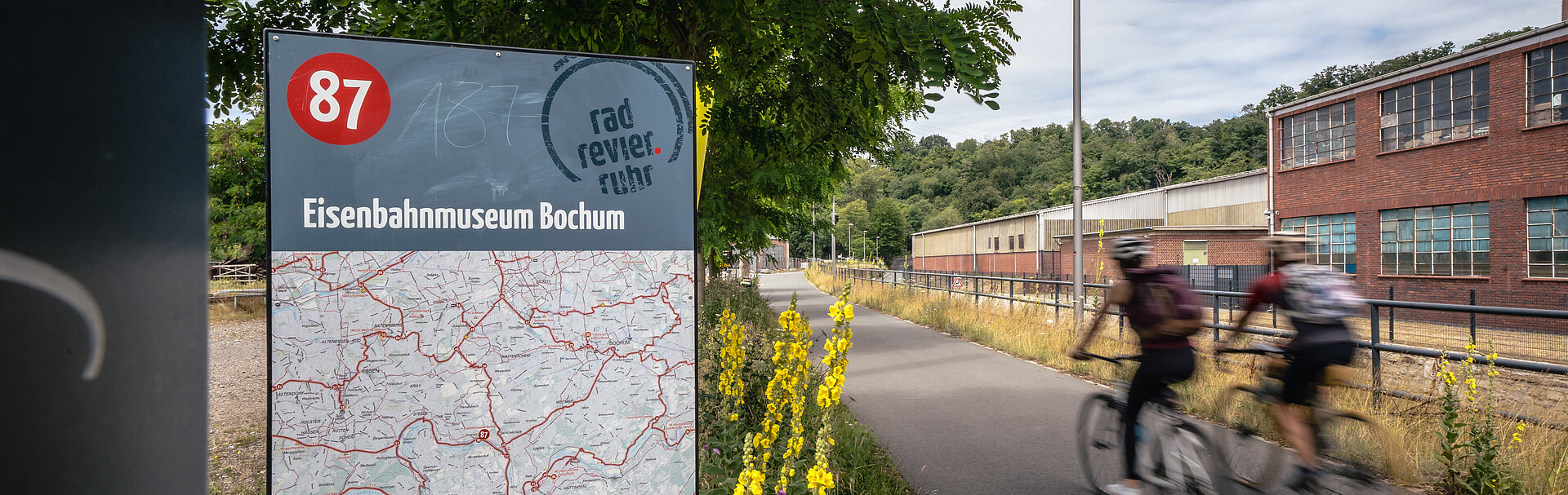 Zwei Radfahrer auf dem Ruhrtal-Radweg auf Höhe des Eisenbahnmuseum Bochum. Im Hintergrund ist das Museum (Industriekultur) zu sehen.