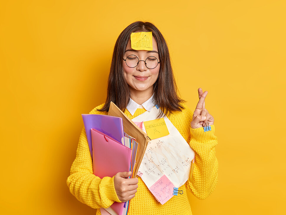 Lächelnde Frau mit geschlossenen Augen, die mit Post-its beklebt ist und den Arm voller Ordner und Schnellhefter hat, kreuzt die Finger ihrer einen Hand im Sinne von "Viel Glück beim Lernen". Der Hintergrund ist gelb.