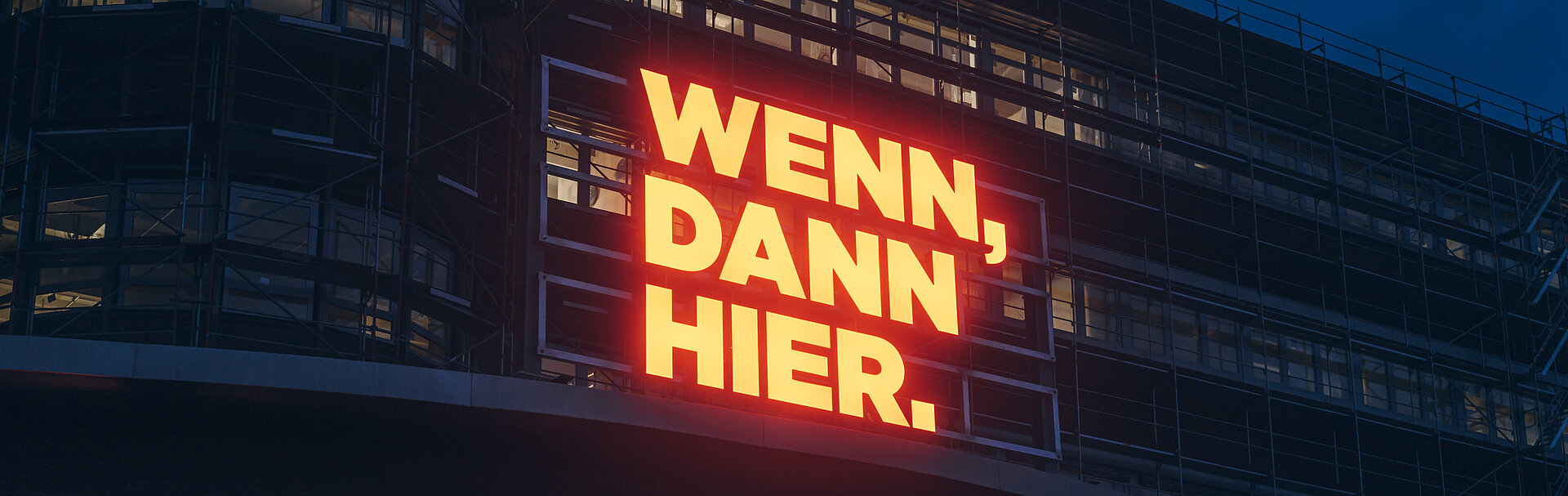 Gelb-leuchtender "WENN-DANN-HIER"-Schriftzug vor nächtlichem Gebäude